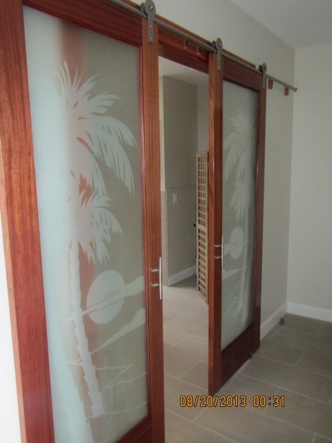 Wood framed interior sliding glass doors with frosted palm tree designs, used as decorative room dividers with warm wood barn door hardware.