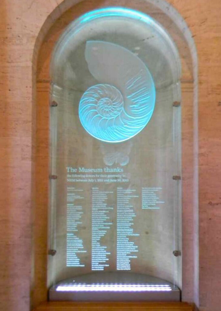 Backlit decorative glass sign with etched nautilus shell and illuminated donor recognition text for wayfinding and display