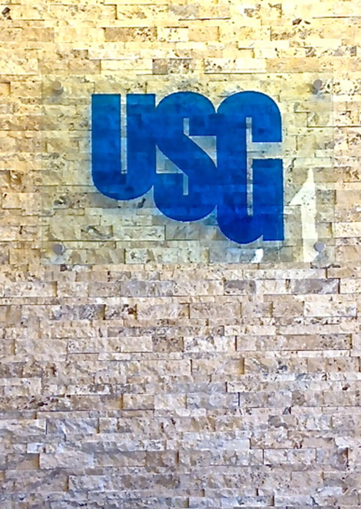 Custom glass wall sign featuring blue USG logo on clear glass panel with mounting hardware, installed on textured stone wall for modern business branding