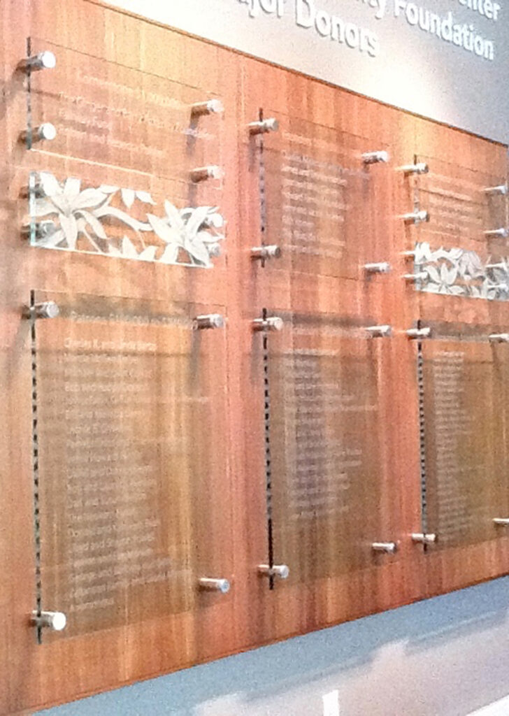 Wall-mounted custom glass signs with etched donor names on multiple glass panels, installed with stand-off mounting hardware for a modern layered look