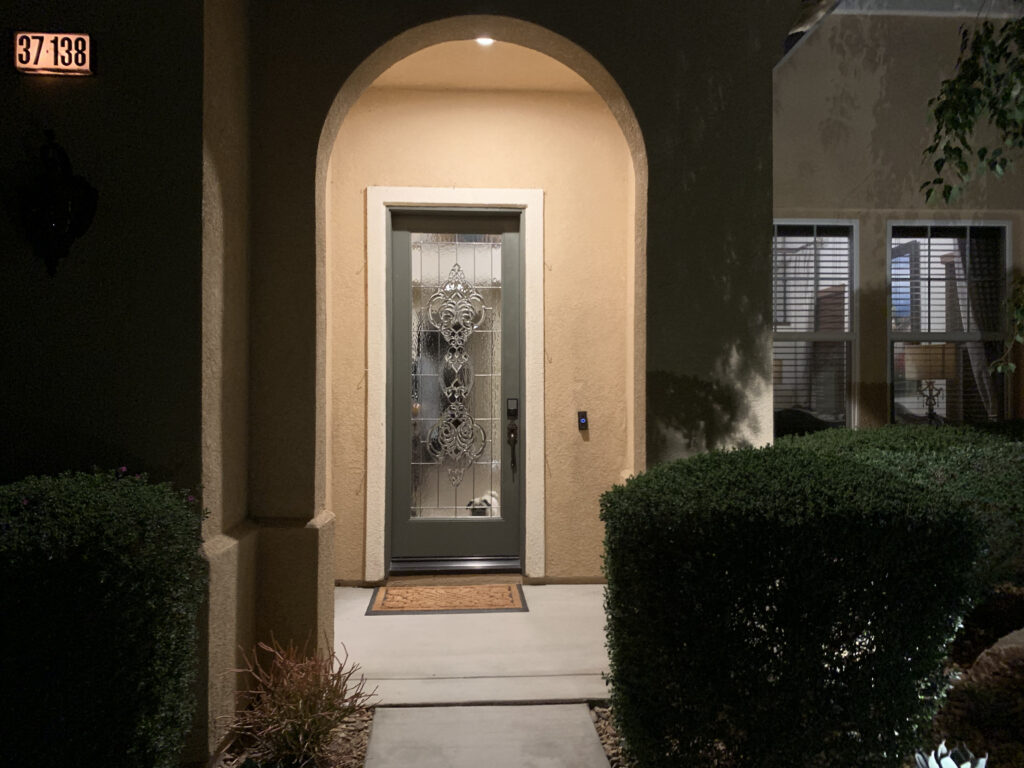 A beautifully designed front door featuring intricate beveled glass, with a cute pug visible through the glass. The scene is softly illuminated by exterior lights, creating a warm and welcoming atmosphere.