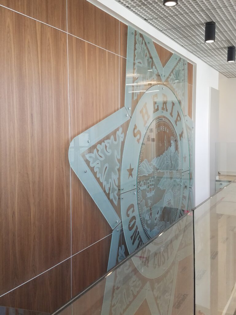 Large custom etched glass wall sign of a sheriff badge mounted with stand-off hardware on wood panel wall, creating a bold professional display