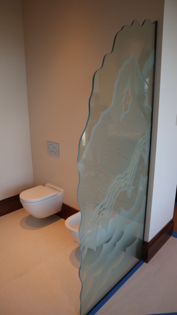 Custom decorative glass partition with carved mountain design used as a private bathroom room divider.