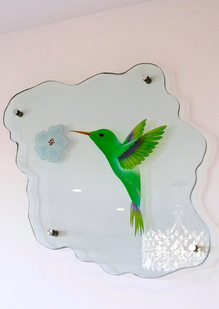 Custom glass sign with painted hummingbird design on shaped glass panel, mounted with stand-off hardware on wall for a sleek decorative display