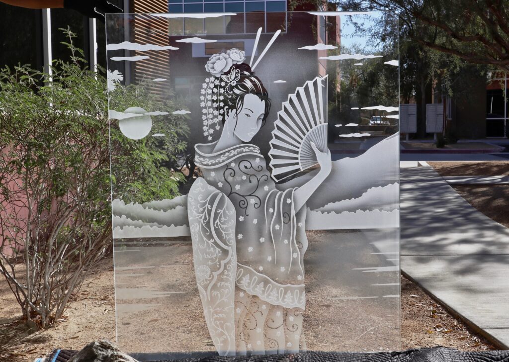Full custom glass panel with geisha artwork etched and carved into clear glass, displayed outdoors to showcase depth, detail, and light interaction