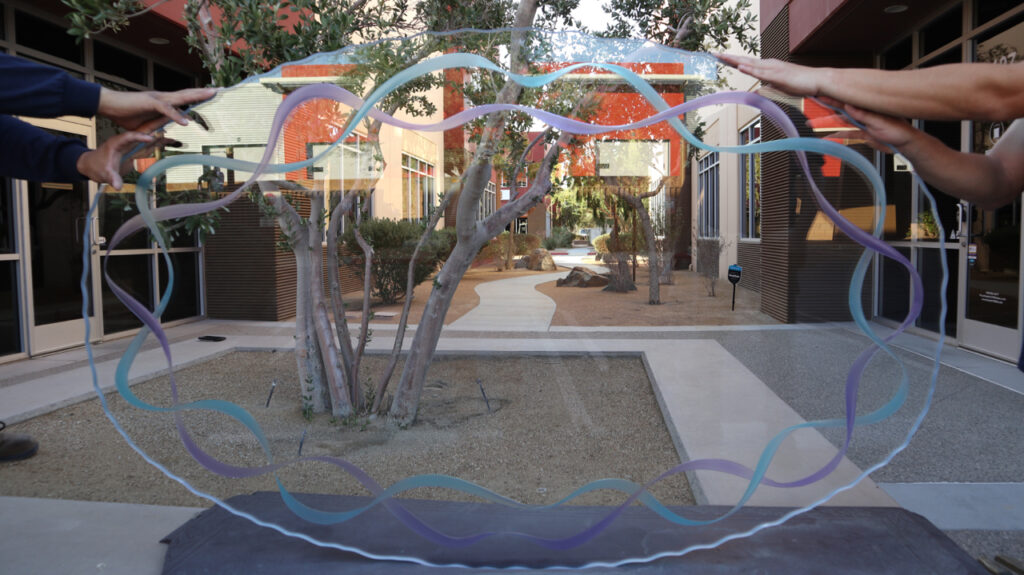 Two hands holding a custom glass table top with colorful, wave-like painted ribbons in a clear glass design. The glass is being positioned outdoors, highlighting its unique and artistic craftsmanship.