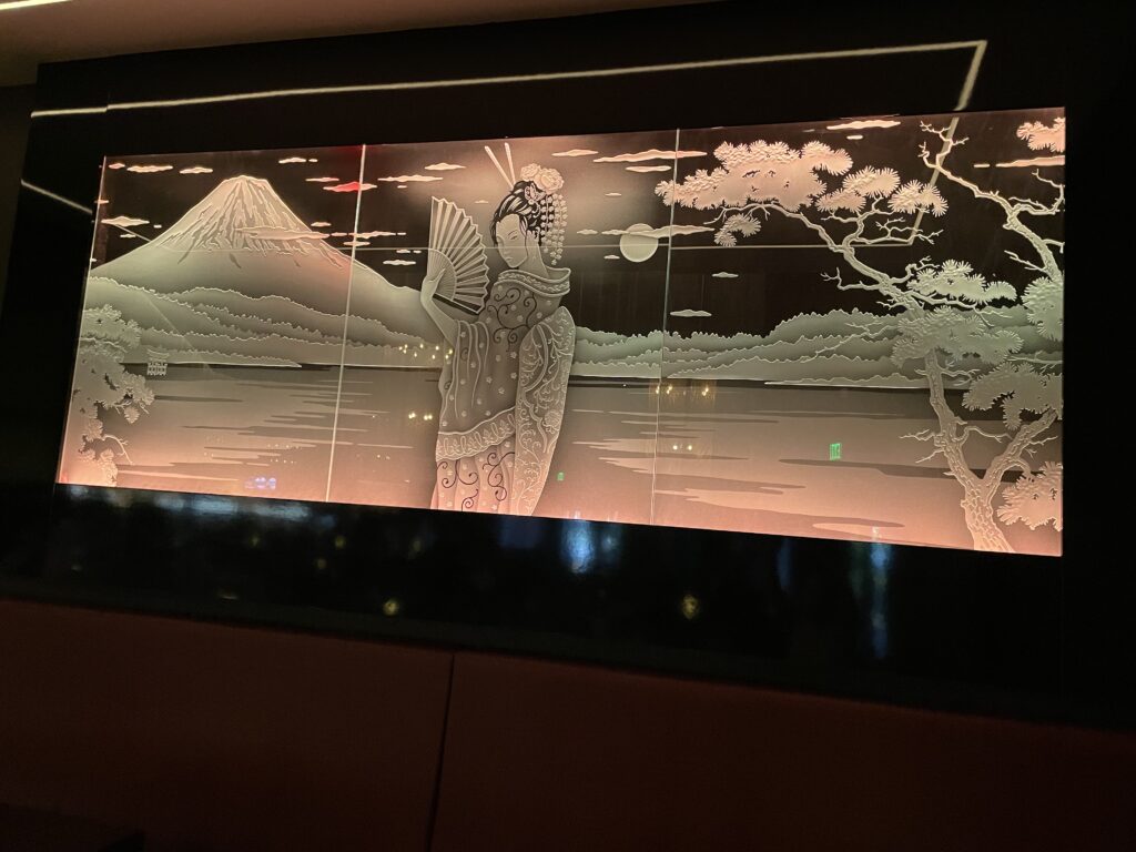 Large multi-panel etched and carved glass wall art with geisha scene, mountain landscape, and trees, illuminated for a stunning architectural display
