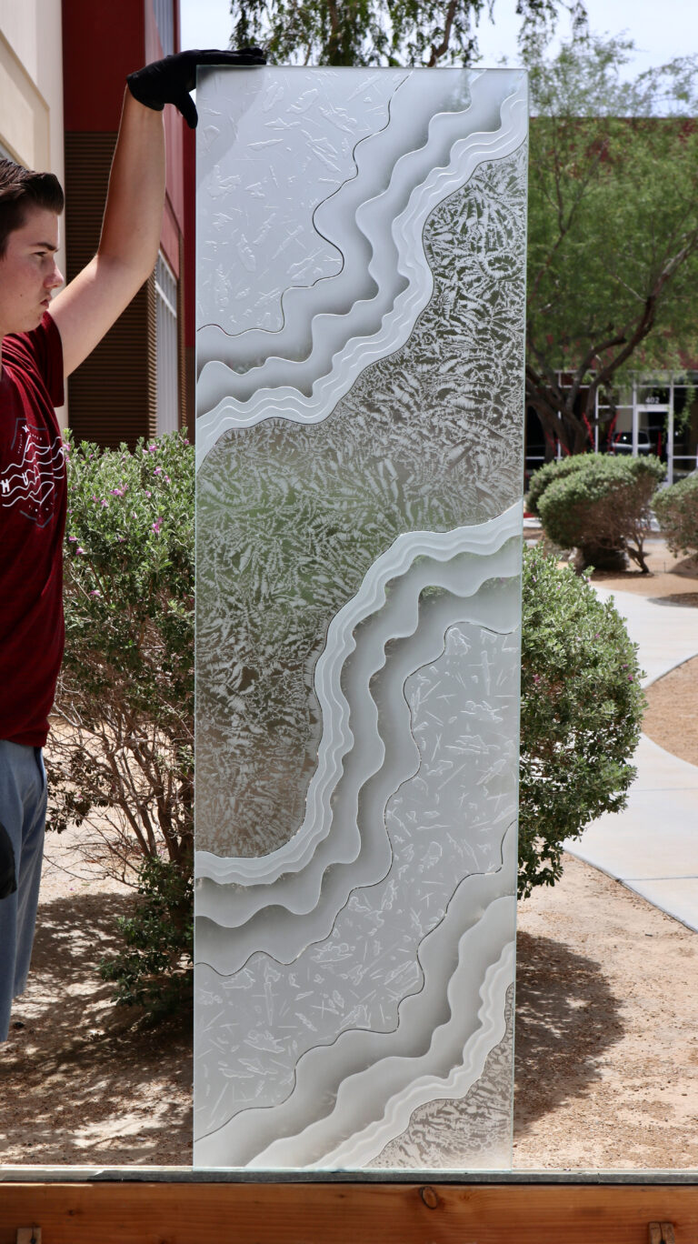 Glass artist holding a tall carved frosted glass panel outdoors, showcasing intricate layered sandblast carving and organic flowing design in custom frosted glass artwork.