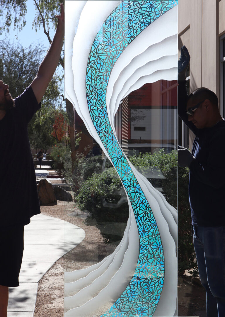 Two glass artisans holding a large carved and painted frosted glass panel, highlighting detailed 3D carved glass design with blue accent pattern and expert craftsmanship.