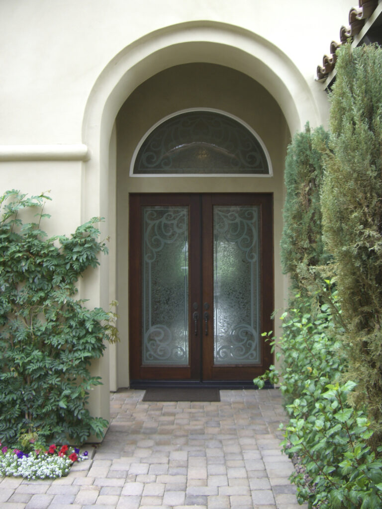 Double front entry doors with frosted glass panels and an arched transom window, set within a stucco exterior surrounded by landscaping.