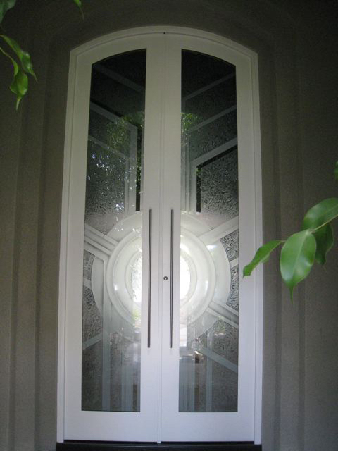 Traditional white double front doors with arched glass panels featuring a bold geometric sunburst design and textured privacy glass.
