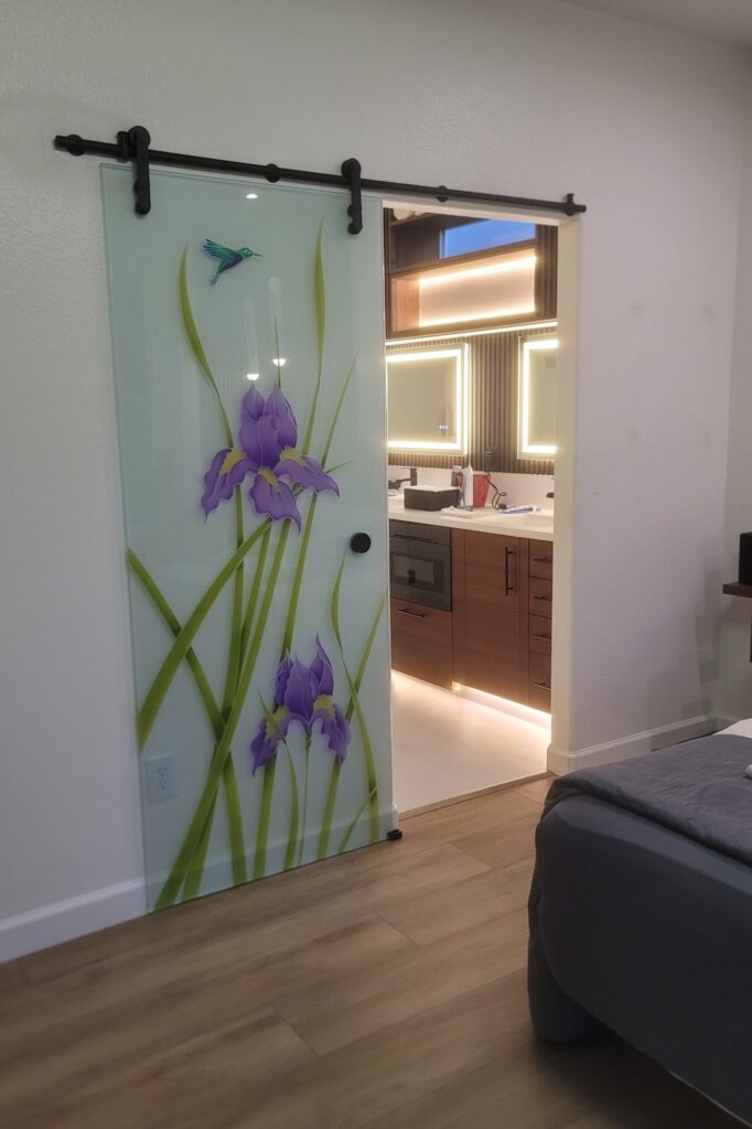 Decorative frosted glass sliding barn door with painted iris flower artwork opening to a modern bathroom with illuminated vanity mirrors