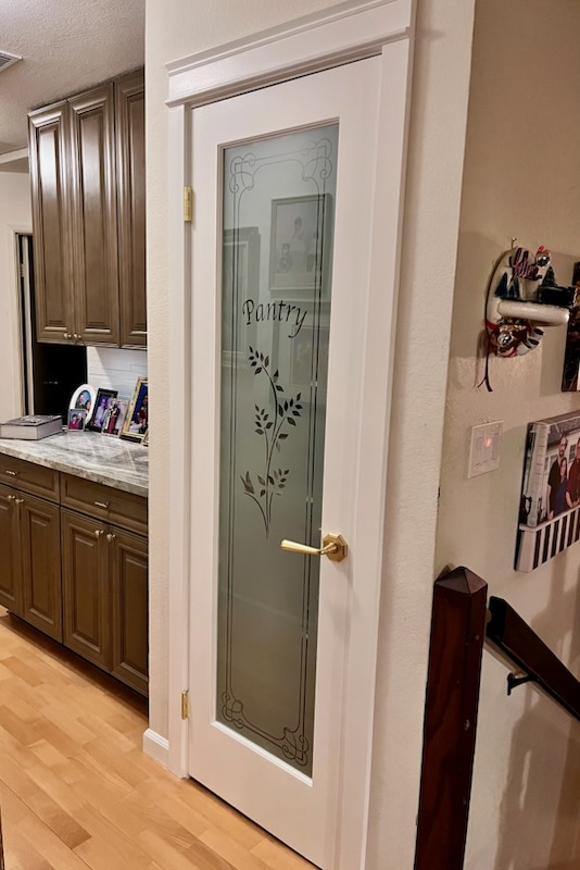 Small kitchen with a frosted glass pantry door featuring the word "Pantry" and a leaf design. The slim hinged door is framed by white trim with a gold handle. Nearby, there are family photos displayed on the wall and dark wood cabinets in the kitchen area. The floor is light wood.