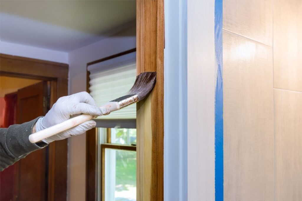 Gloved hand applying wood stain to a door frame using an angled paintbrush.