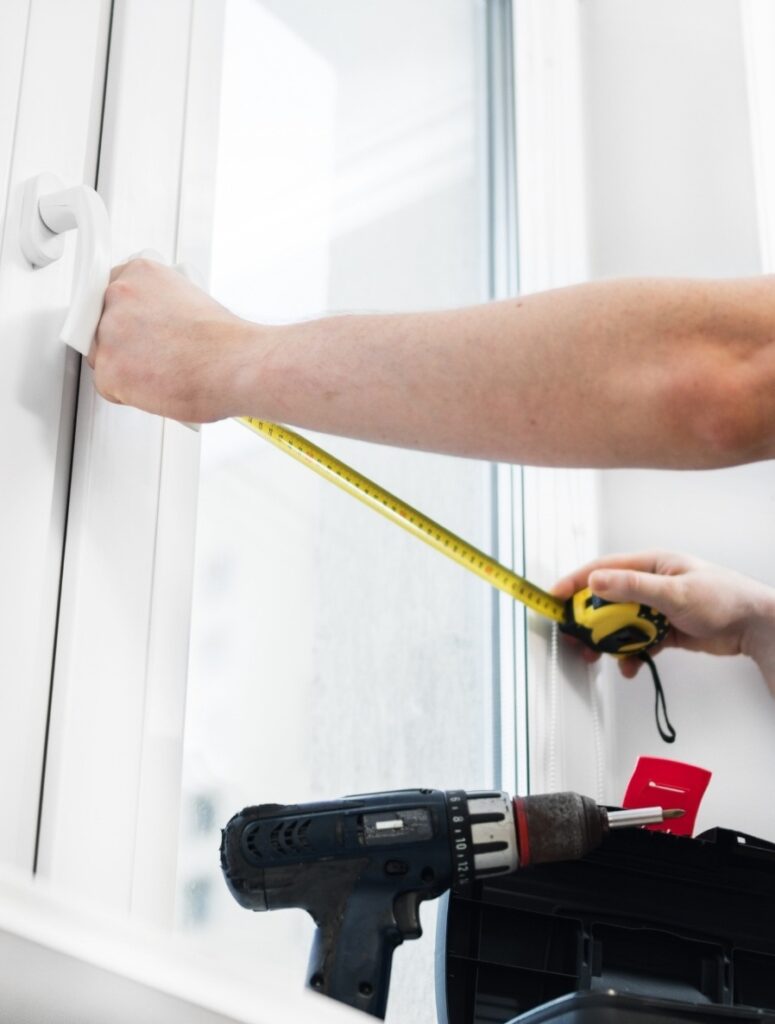 Person measuring a window frame with a tape measure while preparing for installation, with a drill resting nearby &mdash; how to replace a bathroom window.