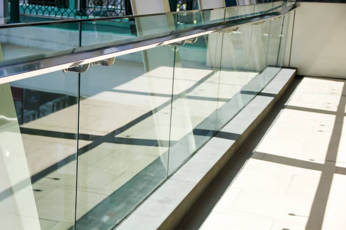 A curved glass balcony railing with a stainless steel handrail is shown in a modern interior space. Sunlight casts a pattern of shadows from the railing's vertical glass panels and the surrounding structure onto the light-colored floor.