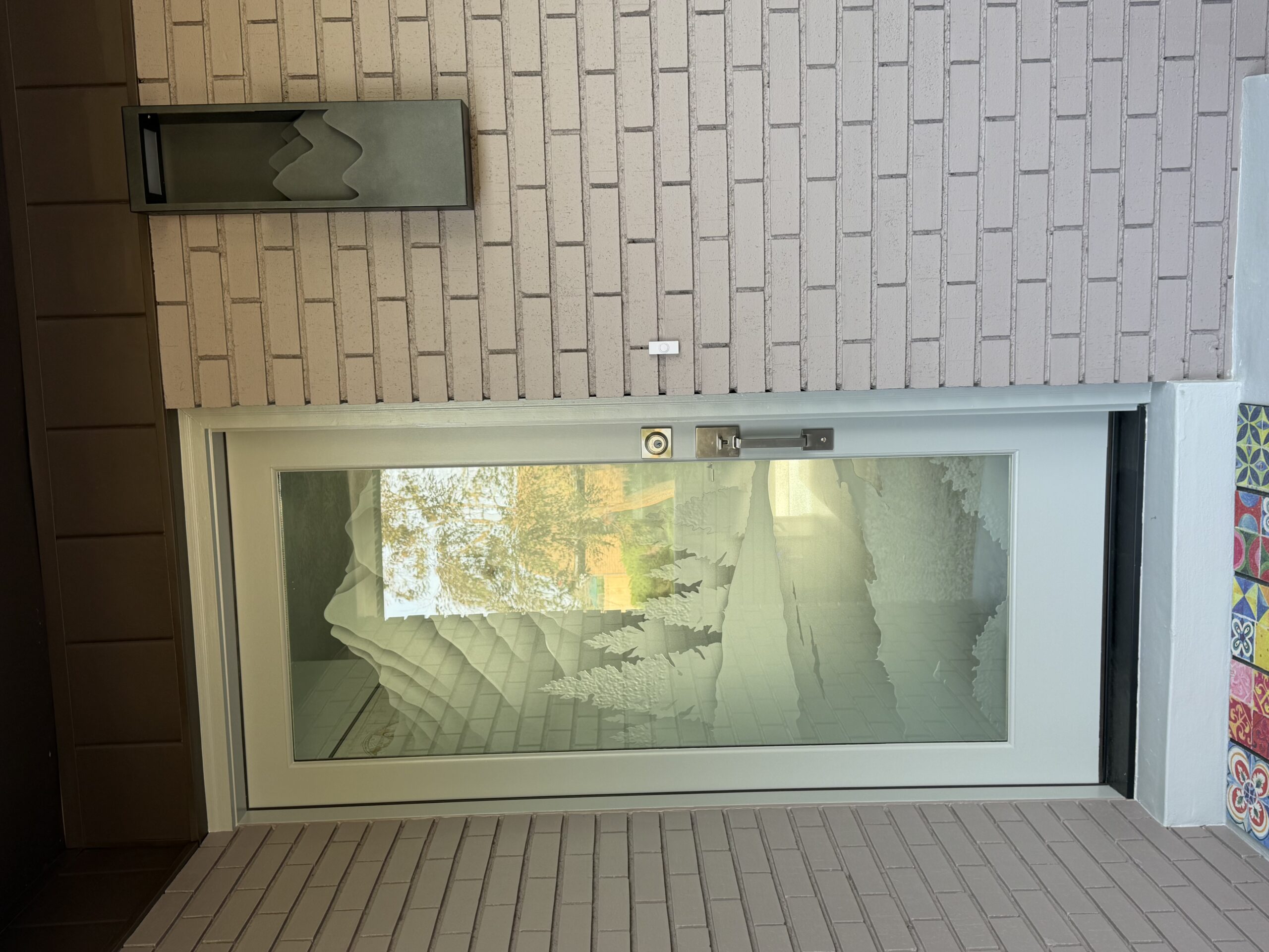Exterior view of a decorative frosted glass front door with a carved mountain and pine tree landscape design, showing how sunlight and reflections shift throughout the day.