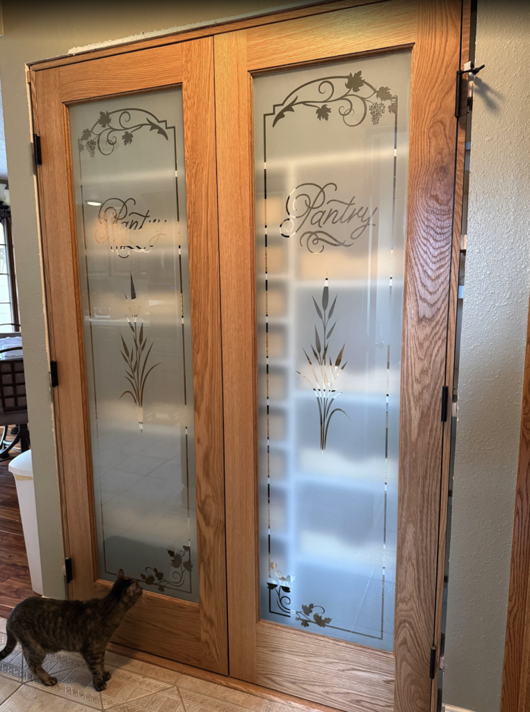 Double glass pantry doors with decorative pantry door glass inserts, etched pantry lettering, cattails, grapevine details, and a cat standing nearby.