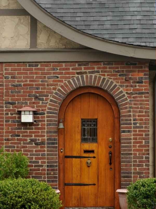 Rustic knotty alder front door with an arched top, black strap hinges, and a small decorative speakeasy glass panel.